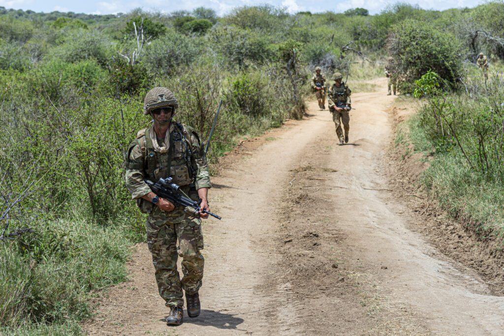 A dirt road with trees to either side takes up most of the foreground. It curves to the right. Three soldiers are visible, walking towards the camera.