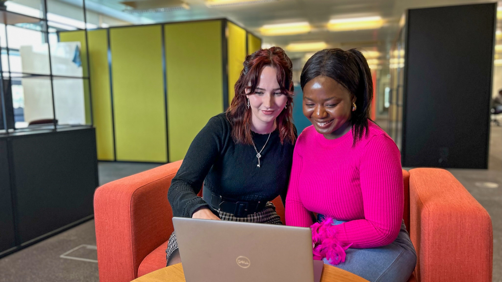 Orla and Olu sit next to each other on an orange sofa. They smile and work together on a laptop computer.
