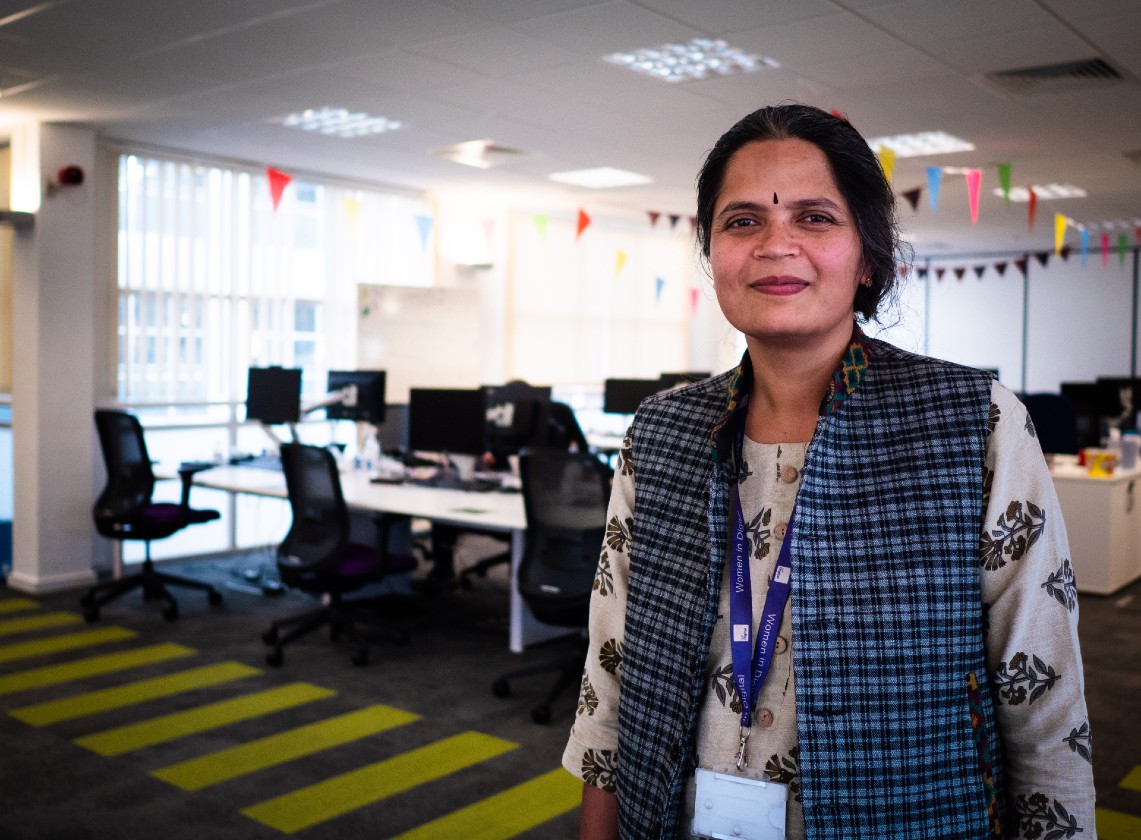 Rashmi stands in an open plan office. She is smiling at the camera.