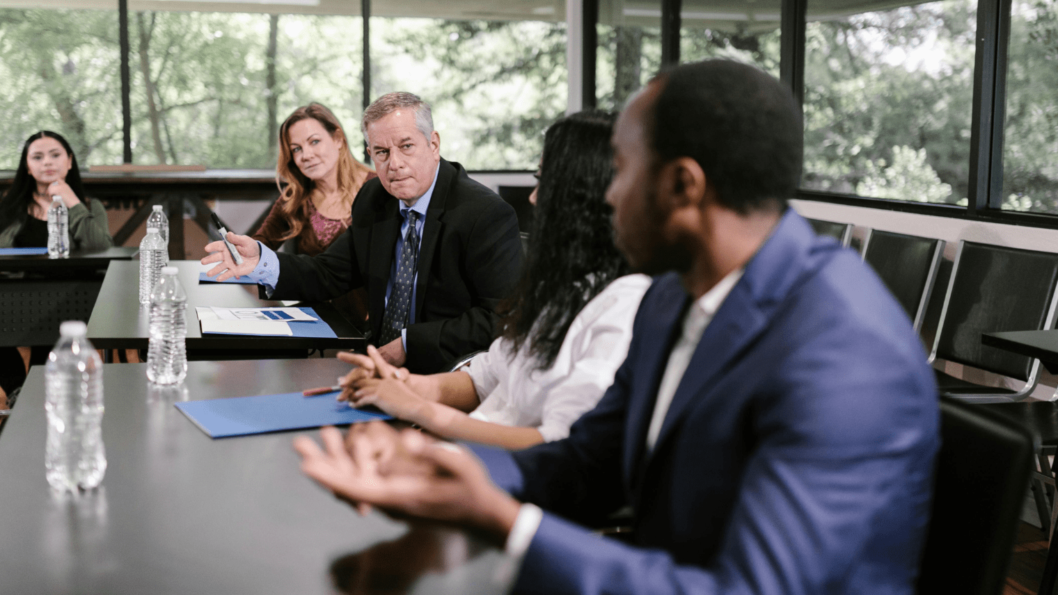 Five people in a meeting room, seated around tables with notebooks, folders, and water bottles as one person speaks and others listen.