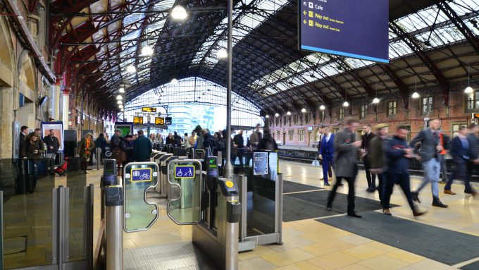 A busy train station platform with people walking and automated ticket barriers in the foreground under a large arched roof