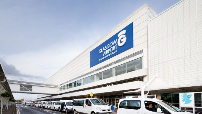 Exterior view of Glasgow Airport terminal building with white taxis lined up in the designated pick-up area, showcasing Glasgow Airport's accessibility features.