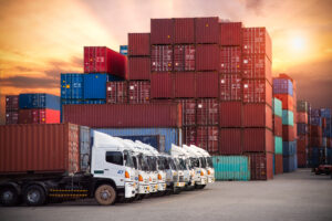 Several white trucks are lined up in front of stacked shipping containers at an industrial location during sunset.