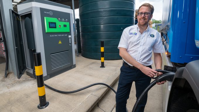 A man in a white shirt and glasses plugs a charging cable into an electric vehicle at a charging station next to a large cylindrical tank.