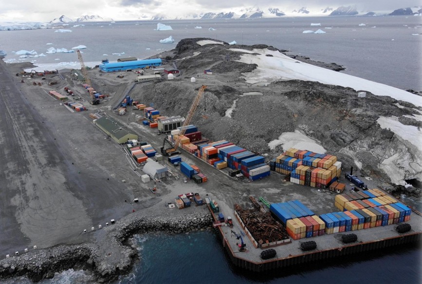A group of people on a wharf near a body of water. There are several shipping containers stacked up and there is a large blue building in the background and ice covered mountains. There are icebergs coming out of the water.