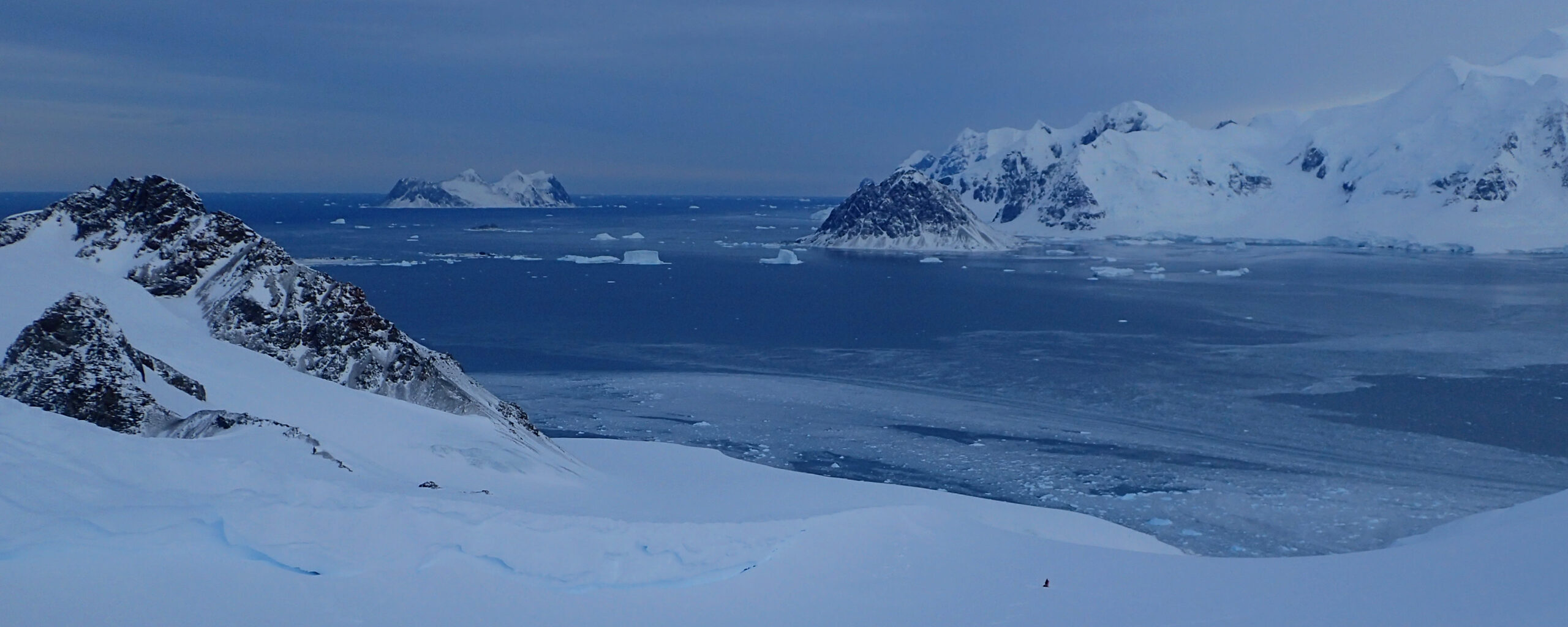 A wide, open Antarctic valley, full of snow and looking down to the sea. Three figures are visible ski treking up the hill in the distance.