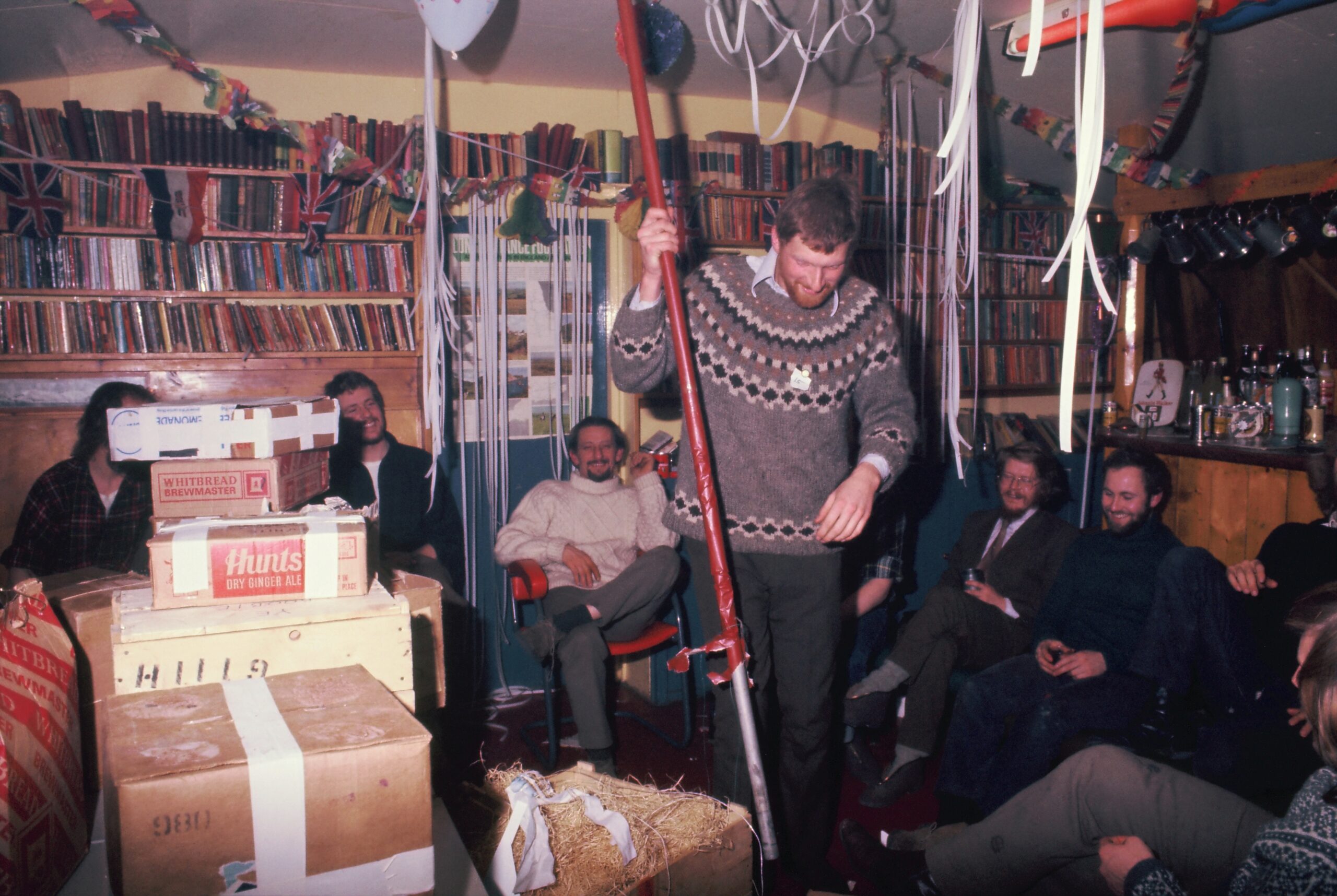 People gather around a pile of wrapped presents as a man unwraps a long cylindrical gift
