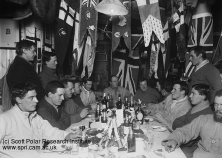 Robert Falcon Scott and his expedition team sit around a table laid with food and decorations. Flags hang from the ceiling featuring Union Jacks.