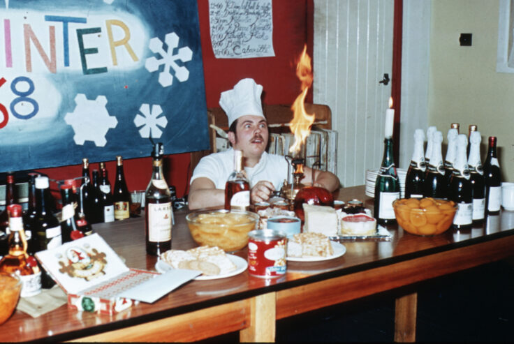 A chef looking surprised as he creates an open flame on a table covered with drinks bottles.