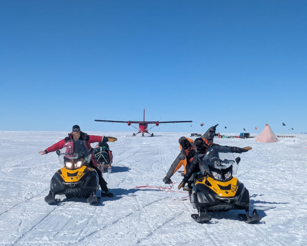 A group of scientists sitting on skidoos in the snow with their arms outstretched imitating the red plane behind them. A blue sky fills the background.