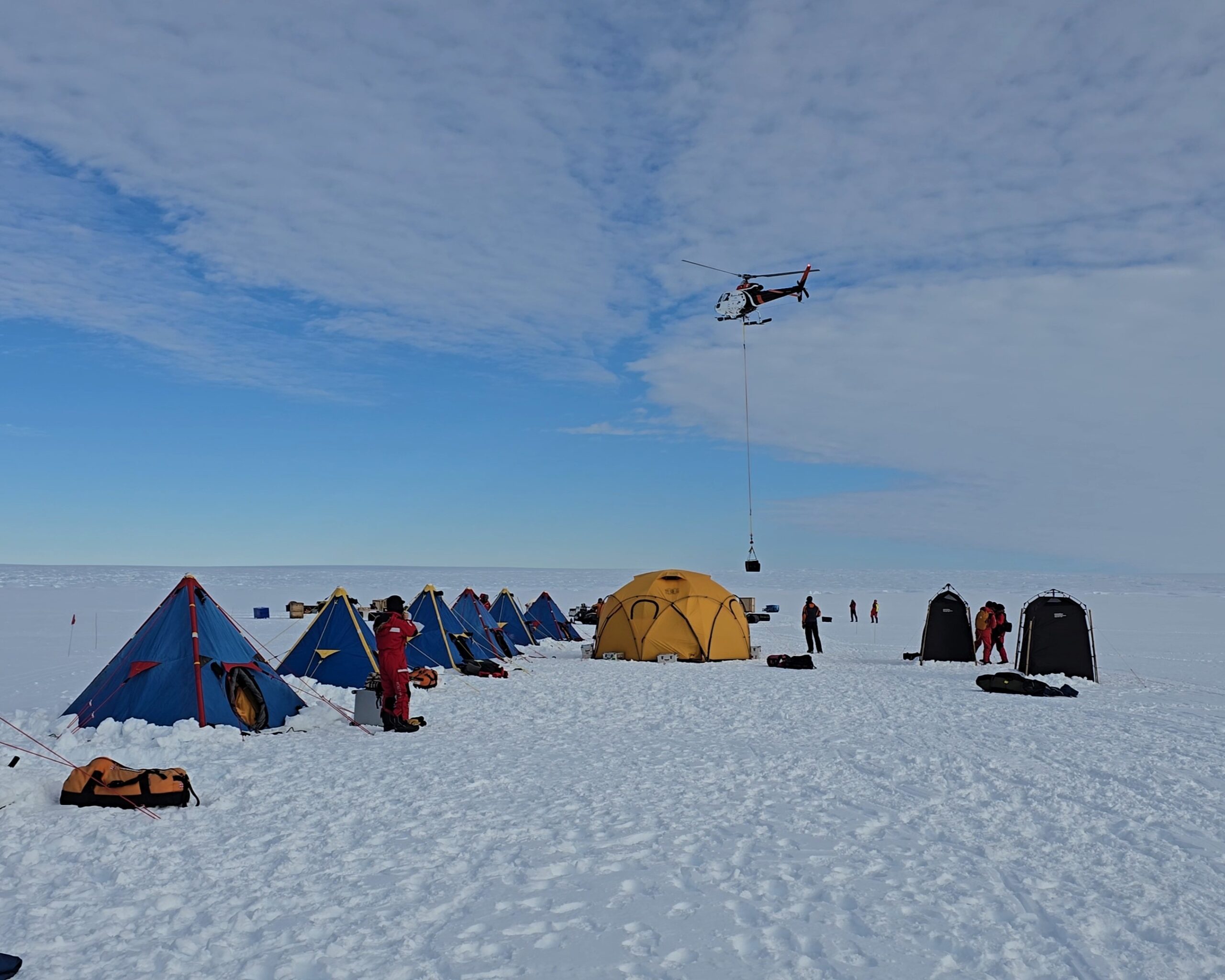 A row of blue and yellow tents on the snows of the Thwaites Glacier. A helicopter delivers supplies in the background.