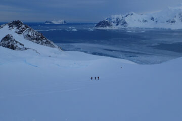 A wide, open Antarctic valley, full of snow and looking down to the sea. Three figures are visible ski treking up the hill in the distance.