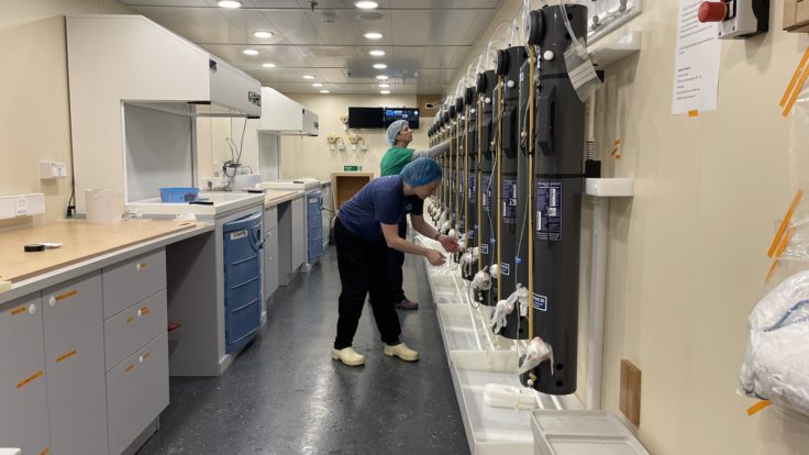 Two scientists wearing scrubs and hair nets work with a series of tanks from the CTD now attached to the wall in a lab.