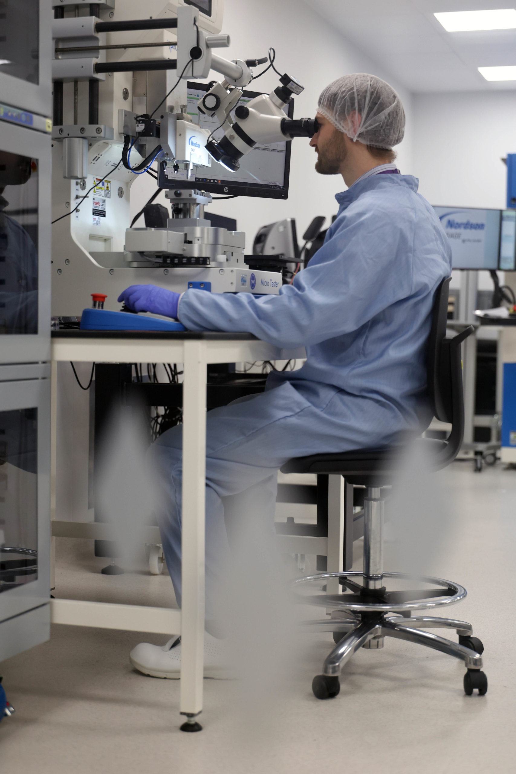 A scientist in protective gear looks into a microscope at a lab workbench with equipment around. - CSA Catapult