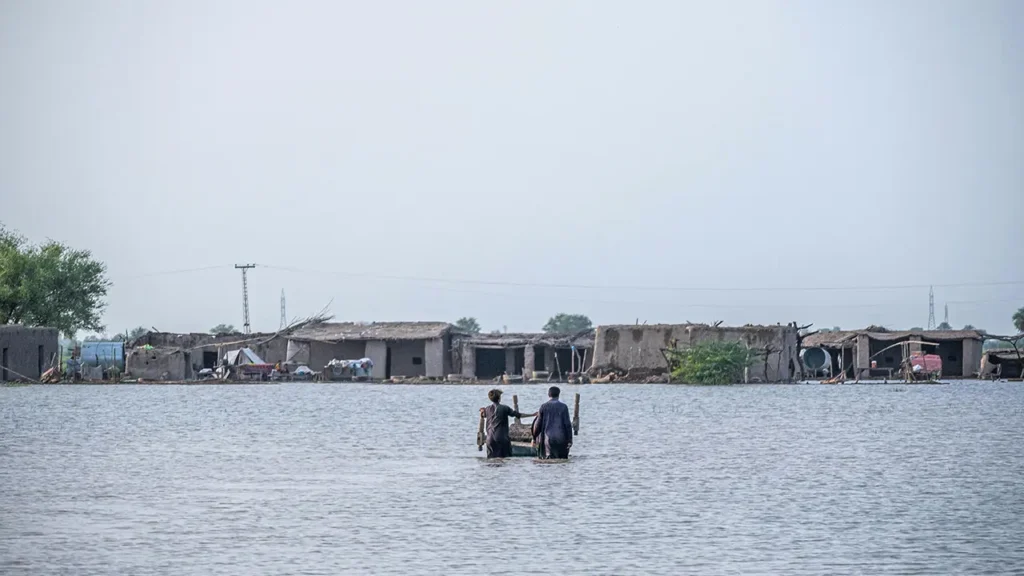 Two people stand wait deep in floodwater, in front of some small buildings and trees