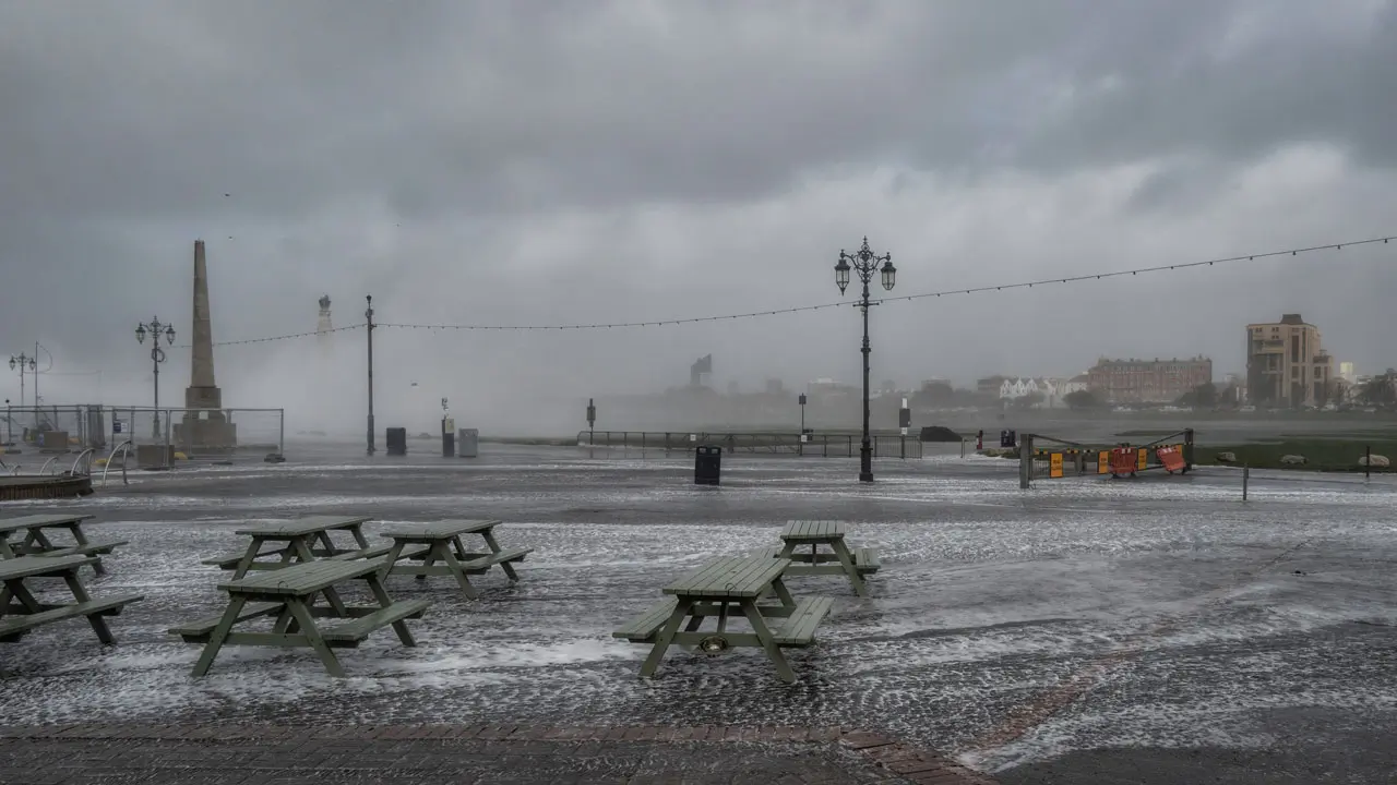 Seafront during storm. Green picnic benches surrounded by water. Grey clouds and seafoam in background.