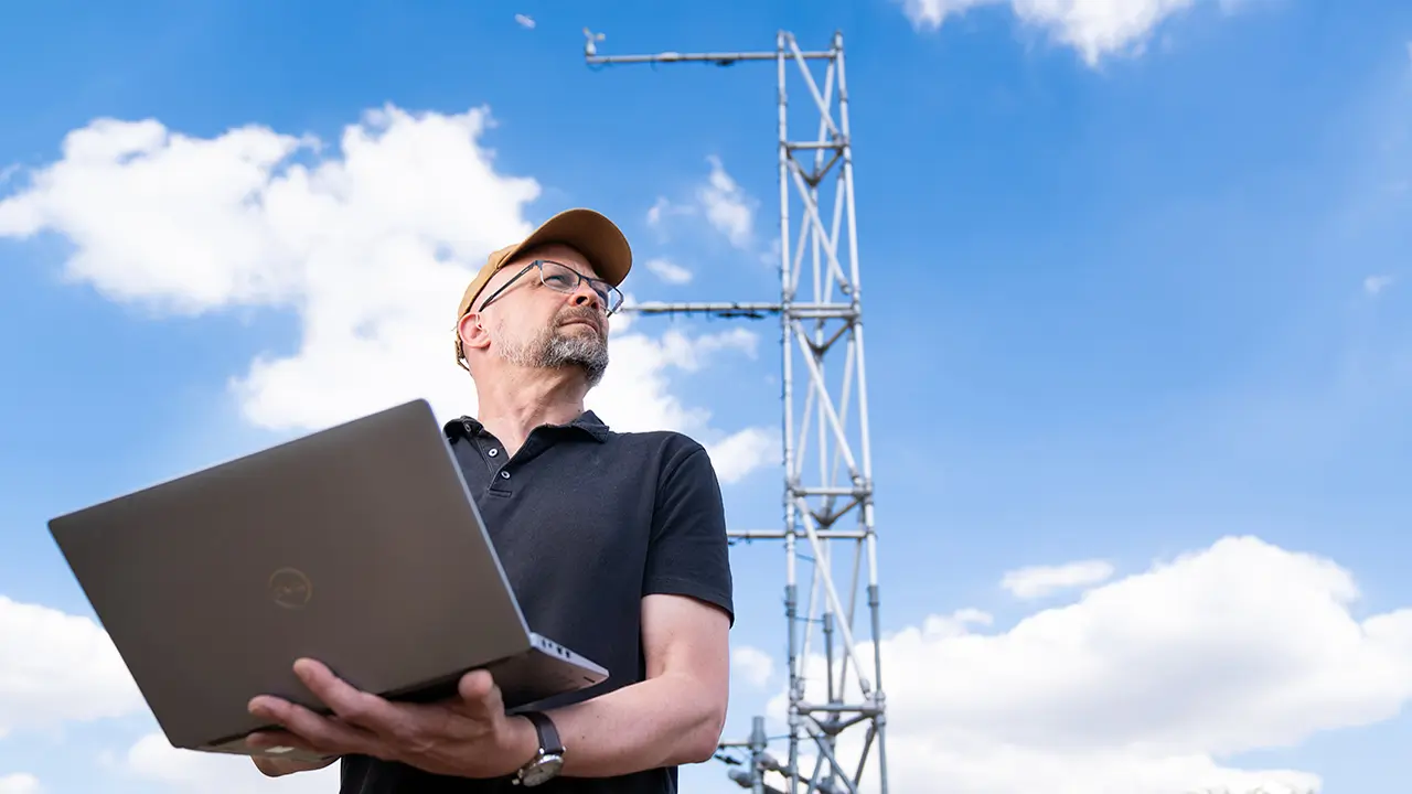 Person wearing glasses and dark blue shirt stands outside looking towards the sky. They stand with a silver laptop in their hands.