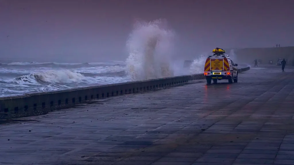 Sea water hitting spraying on coastal wall, with an orange emergency vehicle driving along