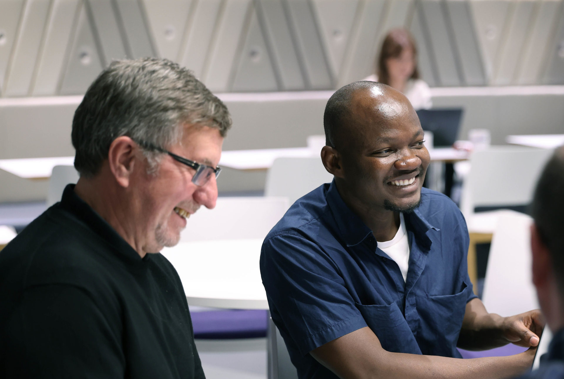 Three men sit at a table, smiling and talking, in a modern, bright classroom with others in the background. - CSA Catapult