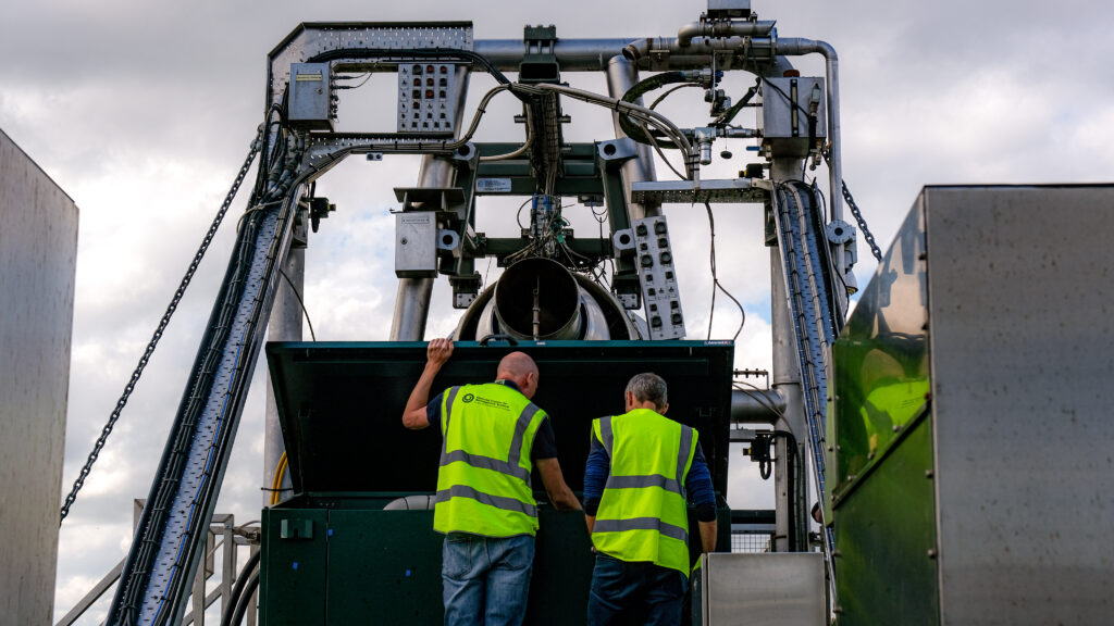 Two people wearing hi vis jackets look into a large metal box, which is in front on an aircraft engine mounted on a large metal structure.