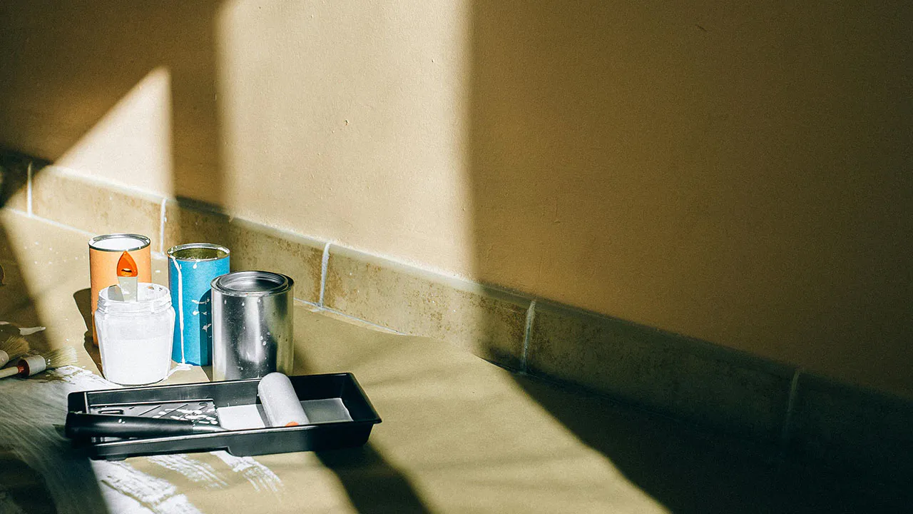 Tins of paint and decorating equipment on the floor, in front of a beige wall. The light from a nearby window is cast onto the wall.