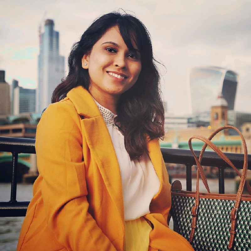 Woman in a yellow coat smiling outdoors with city buildings and a large tote bag in the background.