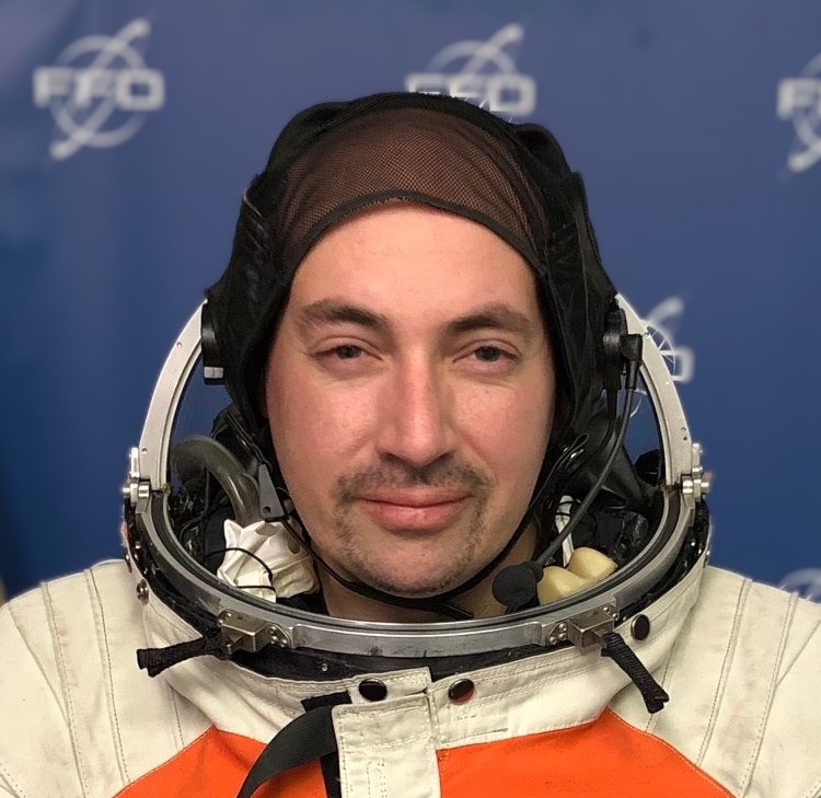 A person in a spacesuit with a headset, posing in front of a blue background with white space logos.