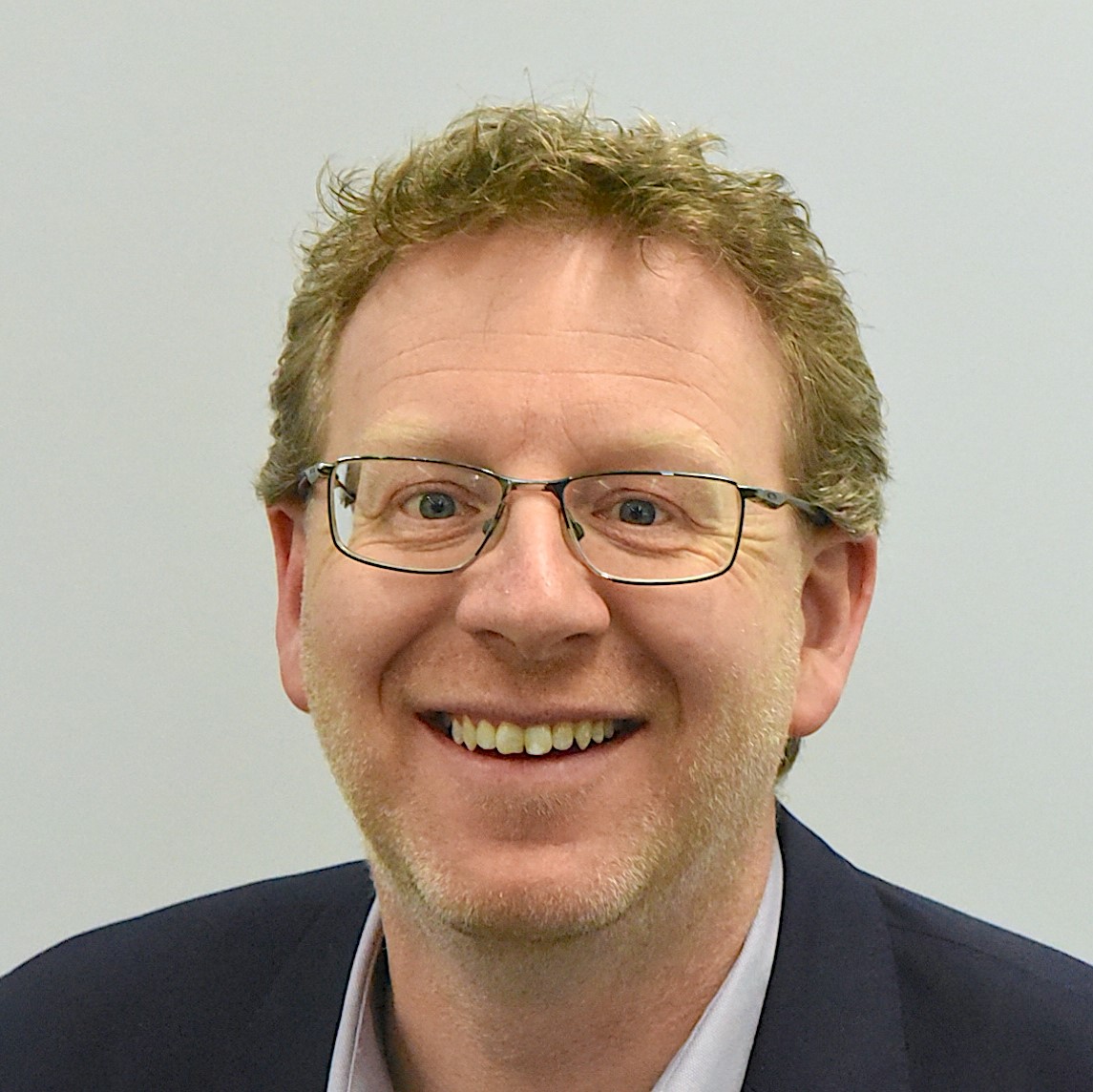 A smiling man with curly light brown hair, glasses, and a blazer against a plain light background.