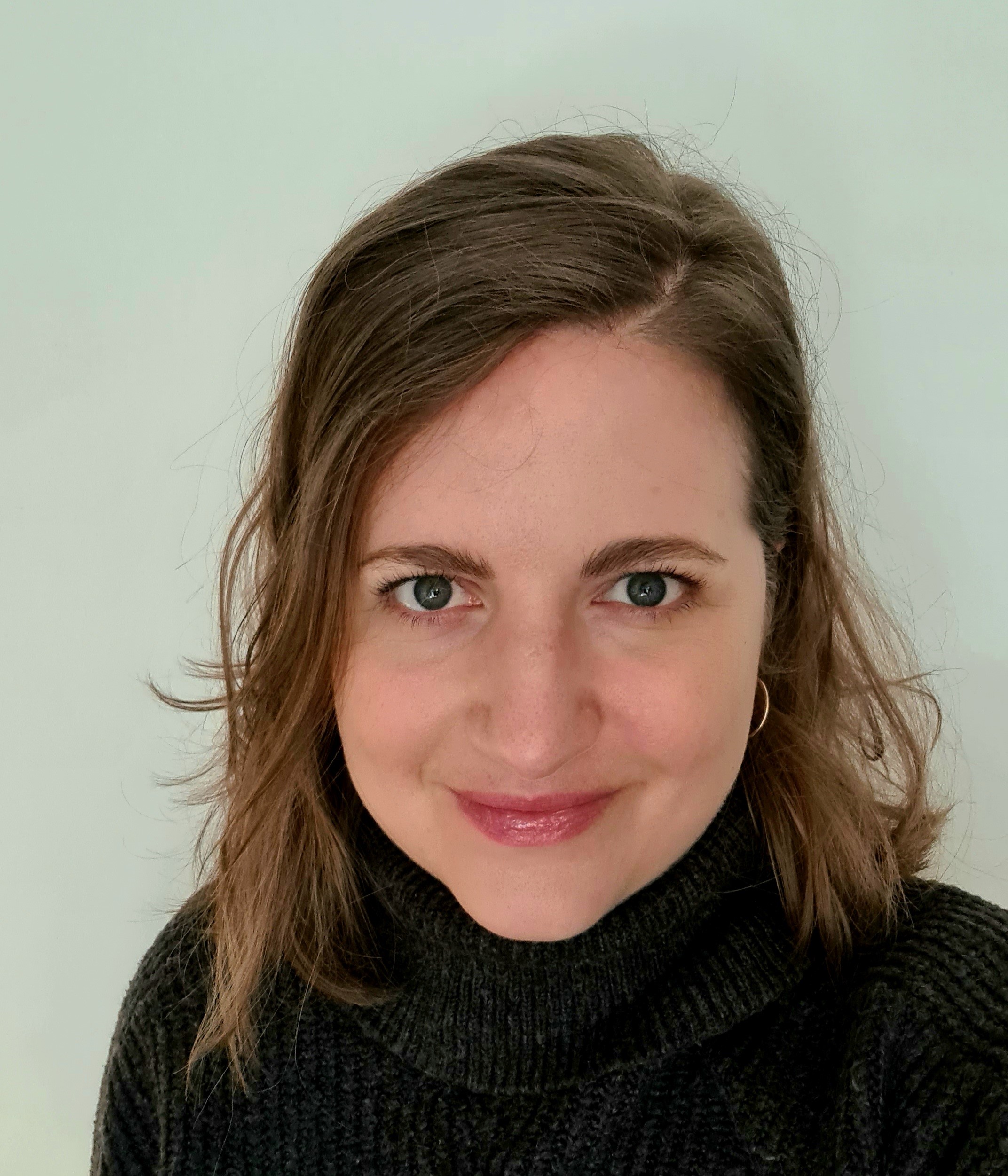 Woman with brown hair and a black turtleneck sweater smiling in front of a plain light background.