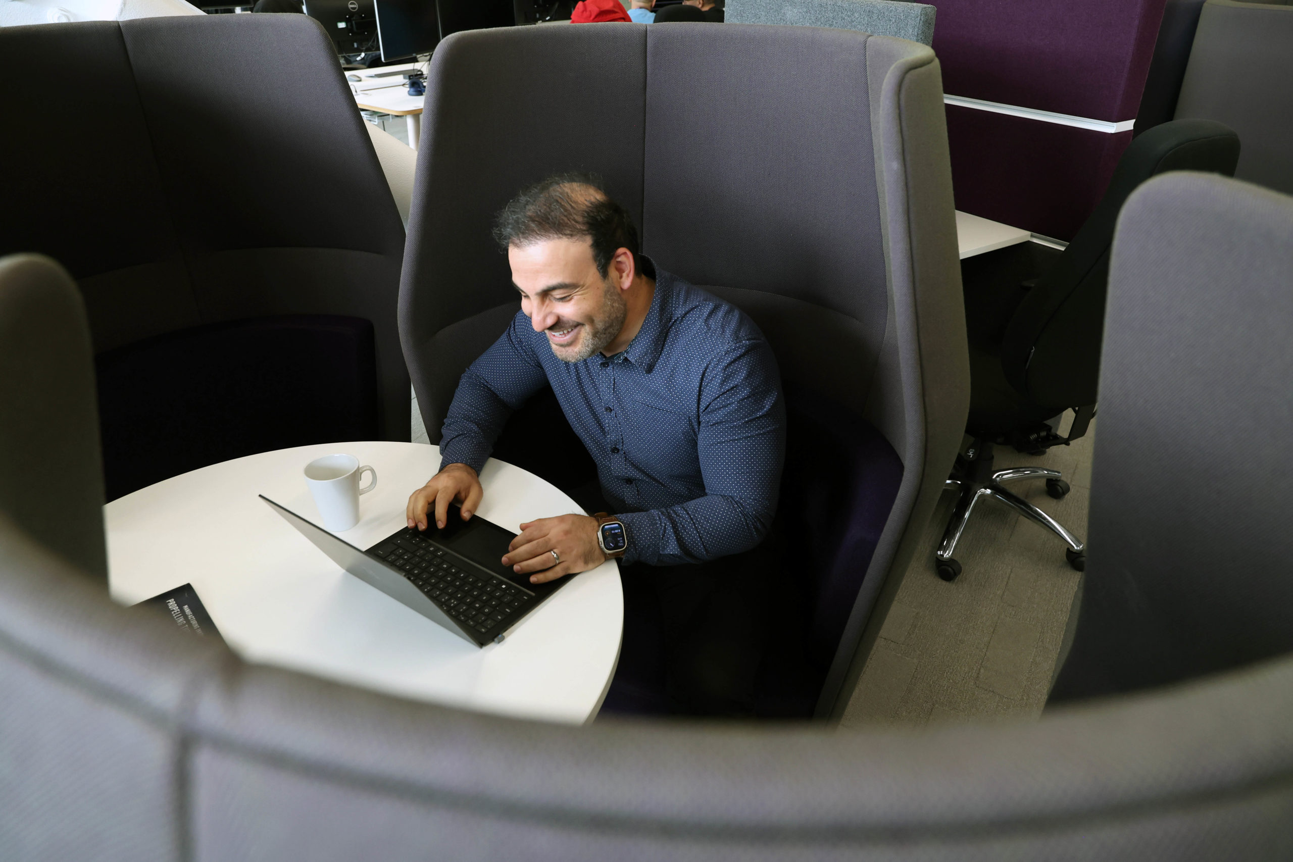 Man smiling while working on a laptop at a round table in a modern office booth, with a coffee mug beside him. - CSA Catapult