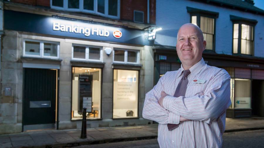 The proud operator of a Post Office branded Banking Hub stands outside the branch in an evening, its lights illuminating the windows. He smiles to camera, arms folded