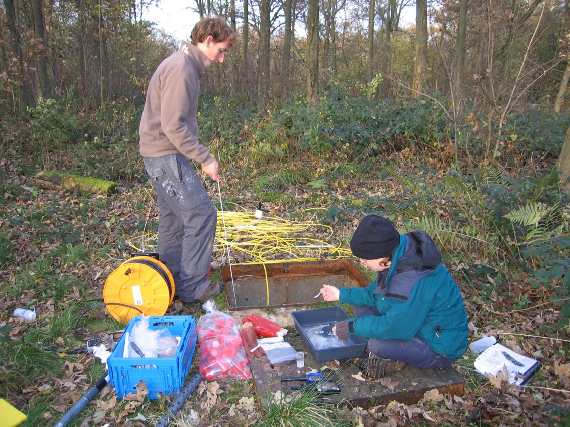 One person standing and one person sitting next to a square borehole entrance, surrounded by sampling equipment, in a wooded area.
