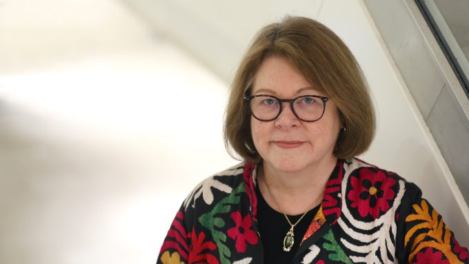 A woman with shoulder-length hair and glasses, wearing a black shirt and a colorful floral-patterned jacket, stands indoors near a staircase, reminiscent of Baroness Brown of Cambridge.