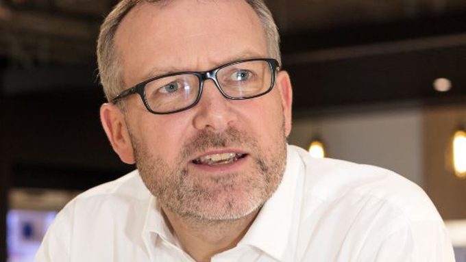 A man with glasses and a beard wearing a white shirt is looking to the side in an indoor setting.