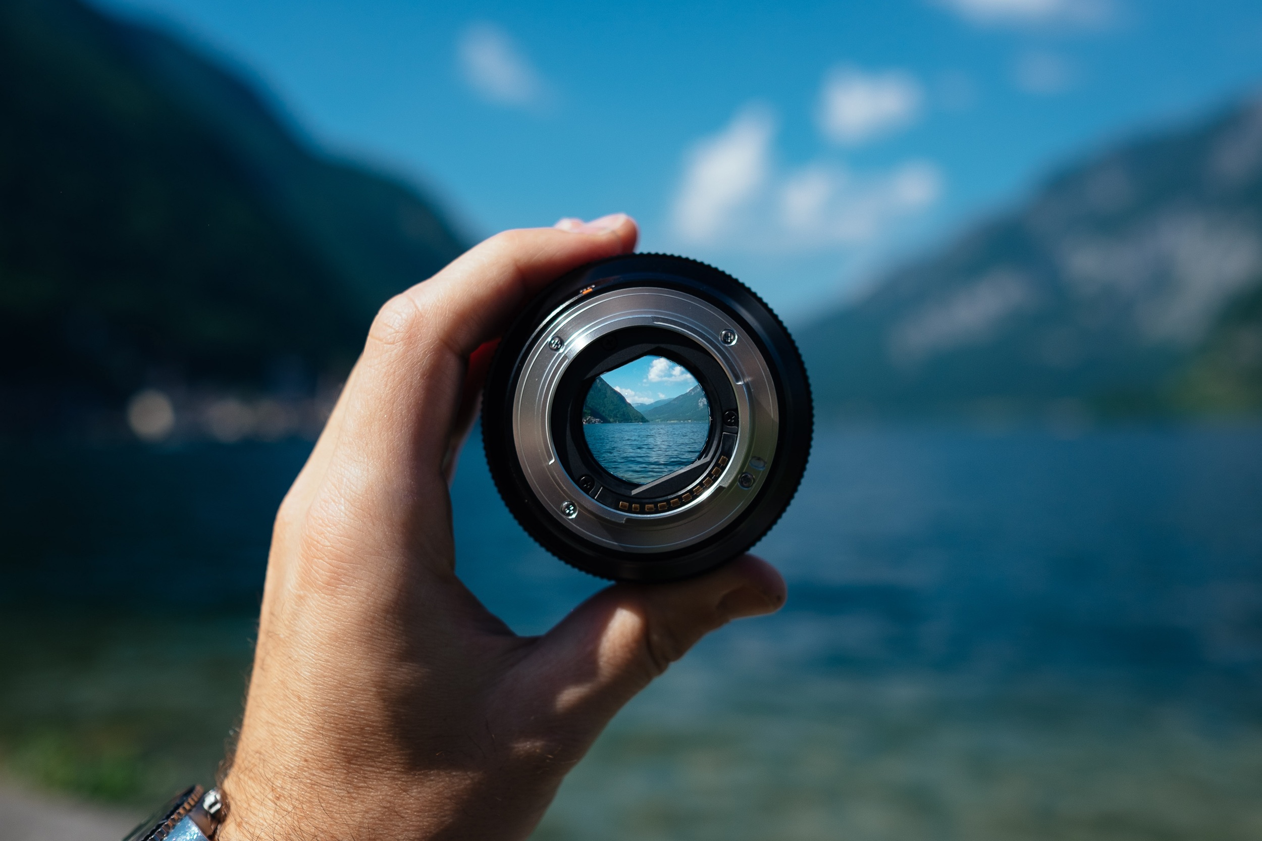 Hand holding a camera lens is shown looking at the far end of a lake on a sunny day. (Credit: Unsplash)