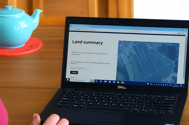 Open Dell laptop on a wooden table displaying a “Land summary” webpage with an aerial map of agricultural land divided into blue outlined plots, with a blue teapot on a red mat beside the laptop.