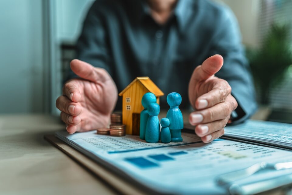 Businessman's hand gestures to protect money, house, wooden dolls represent family. Image represents the concept of asset protection, emphasizing safeguarding financial investments and property