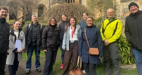 A group of 10 people (mixed gender) stand together smiling outside of an old building with trees and shrubs.