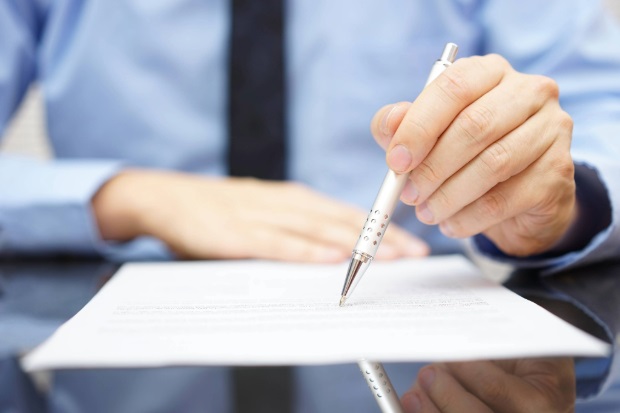 Person sat at a table with a piece of paper laid in front of them with a pen in hand touching the paper