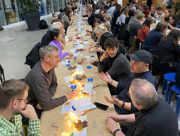 ALT: “People are seated at a long table in an indoor setting, engaged in discussions. The table is decorated with fairy lights, paper notes, and autumnal leaves. The group appears collaborative and focused.”