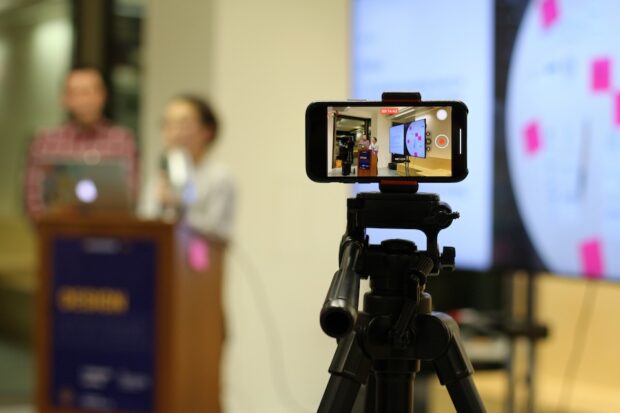 An iPhone capturing a presentation with two people speaking in front of a large screen displaying presentation slides. The presenters are actively engaged, with the slides visible behind them, showing key points of their presentation.