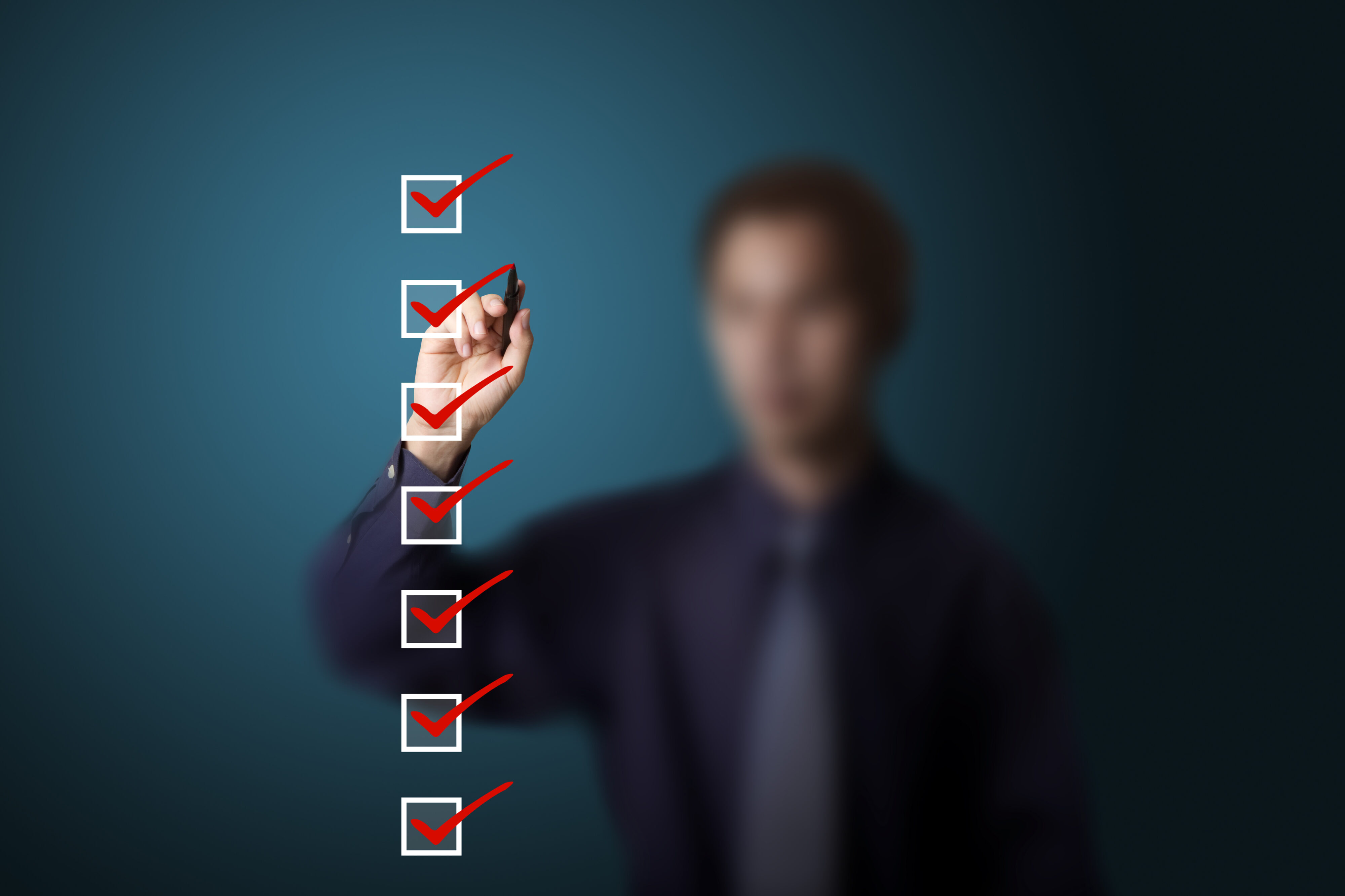 Blurred image of a man in a dark shirt and tie ticking red ticks into white boxes. Credit: Shutterstock