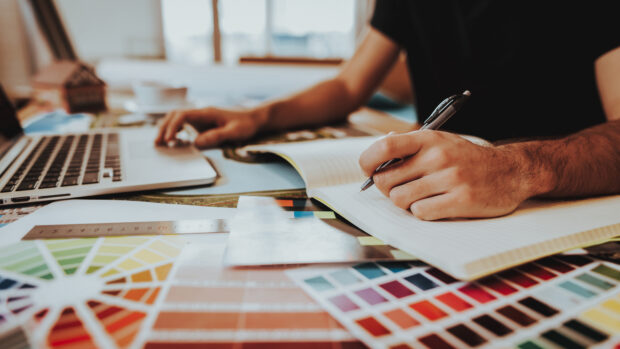A male designer's hnds makes notes in a notebook, whilst using the trackpad of a computer. His desk is covered in colour swatches
