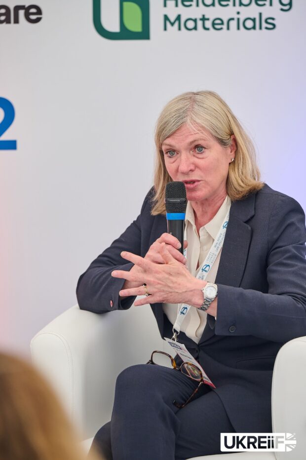 A smartly dressed woman sits in a chair, holding a microphone and addressing a crowd at a conference.