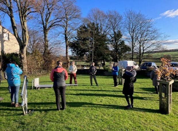 Image shows a small group of young people engaging in nature activities. They are stood outside on a small green, there are trees in the background. A blue sky hangs overhead - the young people are all wearing coats, indicating that it's cold outside and late autumn.