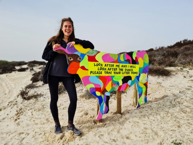 Image shows one of the project leads stood beside an arty cow sign at the dune. The sign is shaped like a cow and is covered in brightly coloured patches of every colour.