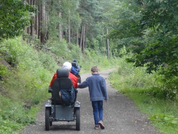 A young children and elderly man in a wheelchair walking down a country lane