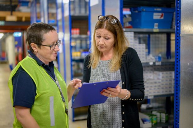 Two women are standing in a warehouse. They are both looking at a clipboard.