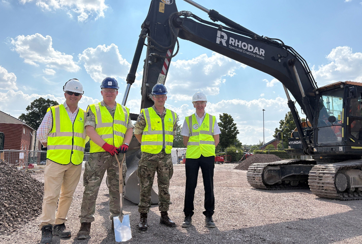 A group of four people in high vis jackets and helmets stand in front of a digger.