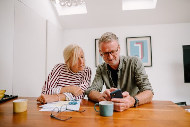 A man and a women sat at a tablelooking at a phone. There are documents and a calculator on the table.